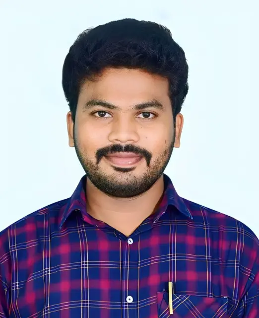 "A young man with short black hair, a well-groomed beard and mustache, smiling slightly while wearing a blue and red checkered shirt against a light background."
