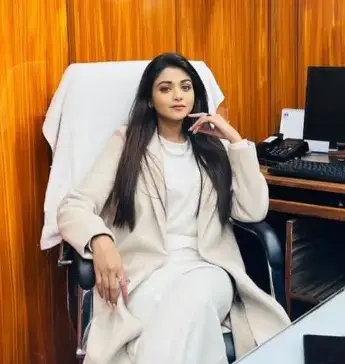 "A young woman with long straight dark hair, wearing a stylish beige coat over a white outfit, sitting confidently in an office chair with her hand near her face."