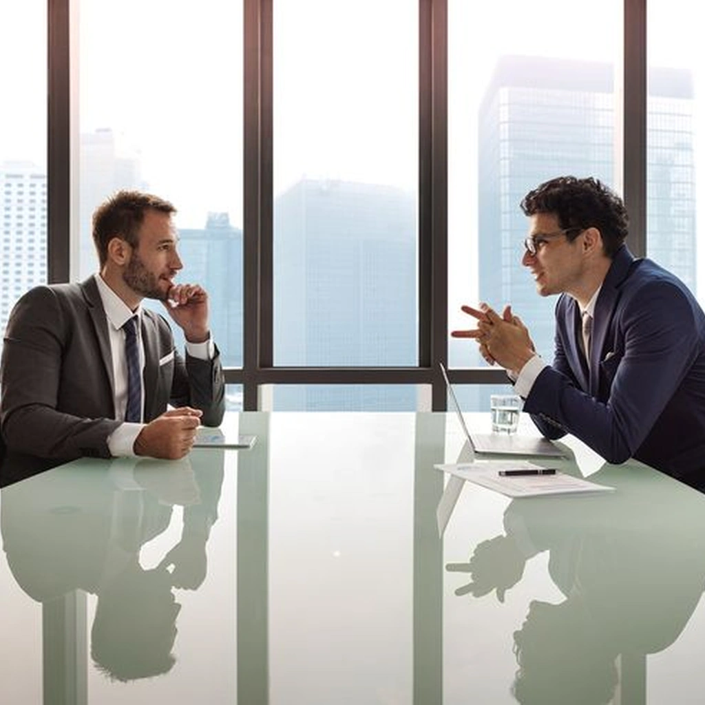 Two corporate executives in suits discussing strategy across a glass conference table in a modern high-rise office, representing client consultation services by a digital marketing company for Dehradun
